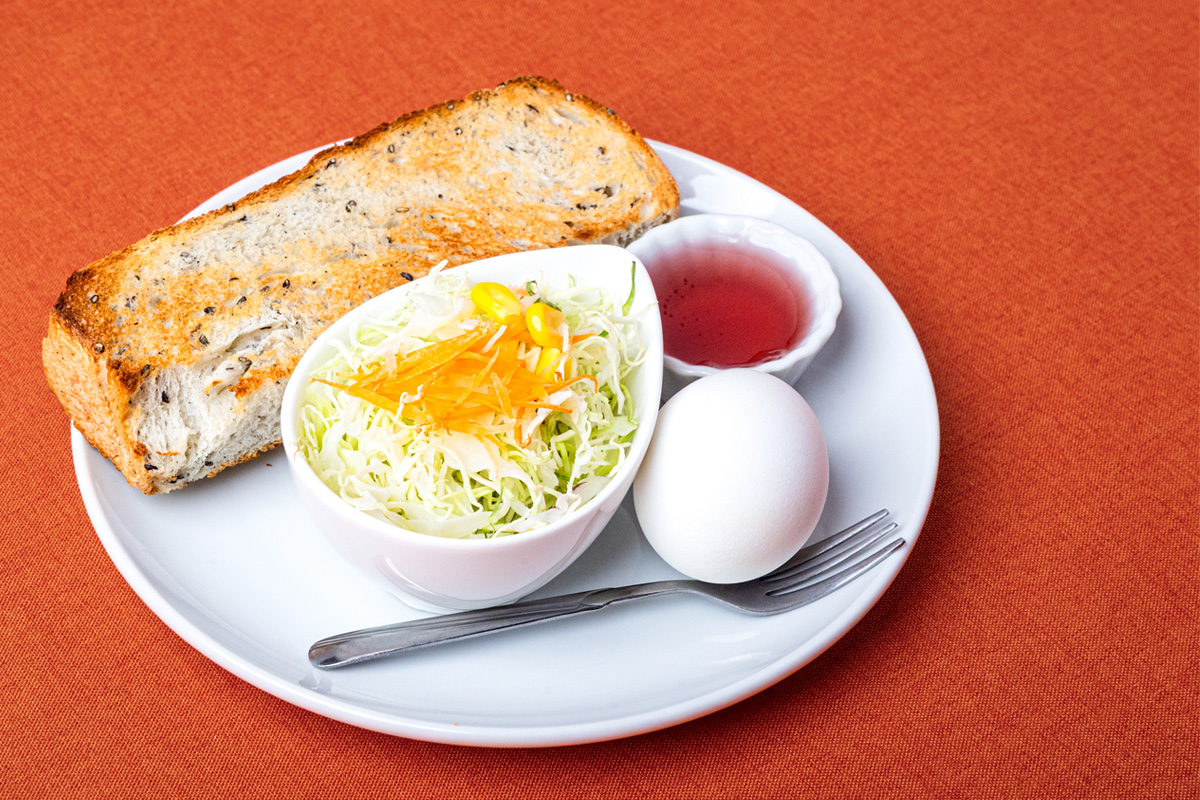 こんがり焼かれた胡麻パン、小皿に盛られたサラダ、ゆで卵、ジャムが白い皿に盛られ、オレンジ色のテーブルクロスの上に置かれている。